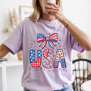 May include: A light purple t-shirt with a patriotic USA design. The design features the word "USA" in red, white, and blue patterns, with a bow and stars. The shirt is a comfortable, casual style.