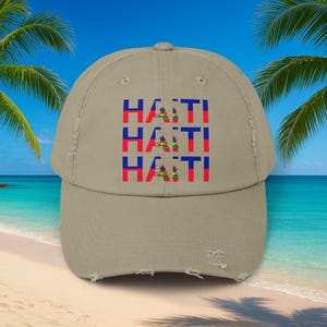 May include: A khaki-colored baseball cap with a distressed, worn look. The front of the cap features the word "HAITI" stacked three times, each in red, blue, and yellow, representing the Haitian flag. The cap has a curved brim.