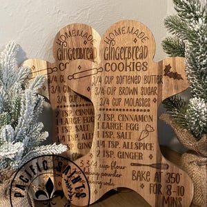 Puede incluir: Tablas de recetas de galletas de jengibre de madera con texto grabado. Las tablas tienen forma de hombre de jengibre y presentan una receta de galletas de jengibre caseras. Se exhibe vegetación festiva con las tablas.