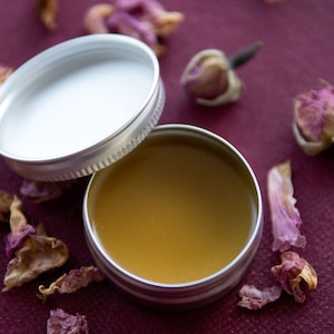 Puede incluir: Una pequeña lata de plata con tapa, llena de un bálsamo amarillo dorado. La lata está sobre una superficie burdeos con pétalos de rosa secos esparcidos a su alrededor.