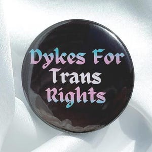 Peut inclure: Un bouton rond noir avec le texte "Dykes For Trans Rights" en lettres roses et bleues. Le bouton est posé sur un fond de tissu blanc et bleu clair, entouré de fleurs roses et violettes.