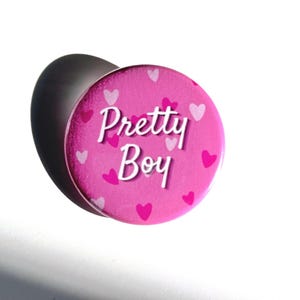 Puede incluir: Botón rosa con corazones blancos y el texto "Pretty Boy" en blanco.