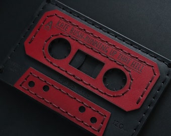 May include: A black leather cassette tape wallet with red stitching. The text "KATE BUSH - RUNNING UP THAT HILL" is printed on the front.