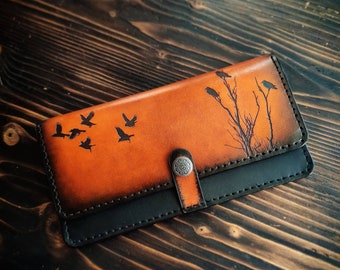 May include: A brown leather wallet with a black flap and a silver button closure. The wallet has a design of birds flying and a tree with branches on the front.