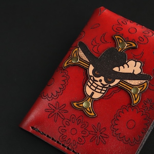 Puede incluir: Cartera de cuero roja con un diseño de calavera y huesos cruzados. La calavera lleva un sombrero de vaquero negro y un pañuelo amarillo. La cartera tiene un patrón floral.