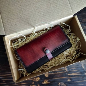 May include: A burgundy leather wallet with a black leather trim and a silver button closure. The wallet is in a box with shredded paper.