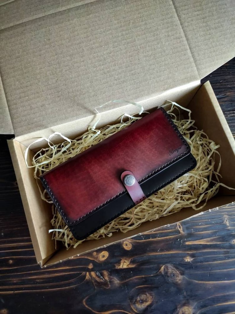 May include: A burgundy leather wallet with a black leather trim and a silver button closure. The wallet is in a box with shredded paper.
