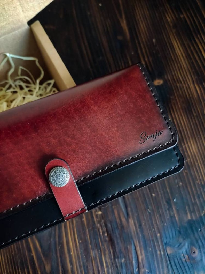 May include: A burgundy leather wallet with a black leather trim and a silver snap closure. The wallet has the name "Sonja" embossed on the front.