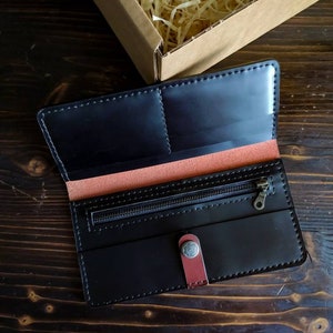 May include: A black leather wallet with a zippered pocket and a snap closure. The wallet has a red leather accent and is open to reveal the interior.