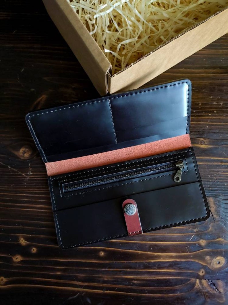 May include: A black leather wallet with a zippered pocket and a snap closure. The wallet has a red leather accent and is open to reveal the interior.