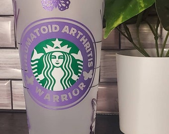 May include: A white reusable cup with a purple and green design. The cup features a Starbucks logo with the text "Rheumatoid Arthritis Warrior" around it. There are purple butterflies on the cup.