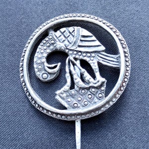 May include: A silver-toned pin featuring an ornate design of a stylized bird within a circular frame. The bird has detailed patterns and a long, curved beak. The pin has a long, pointed needle for attachment.