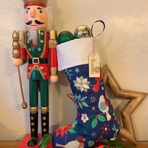 May include: A festive Christmas stocking with a blue floral design, filled with baubles, beside a wooden nutcracker and a wooden star. The stocking has a white cuff and a wooden tag that says "Namaste".