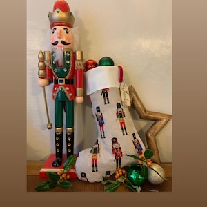 May include: A wooden nutcracker soldier with a red and green uniform stands next to a Christmas stocking. The stocking is beige with a pattern of nutcracker figures. Ornaments and a star-shaped decoration complete the festive scene.