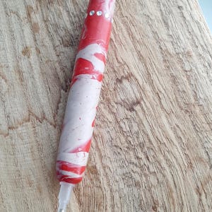 Könnte beinhalten: Ein roter und weißer Spiral-Stift mit klaren Strasssteinen auf der Oberseite. Der Stift liegt auf einer Holzoberfläche.