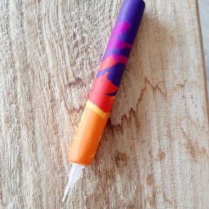 Könnte beinhalten: Ein farbenfrohes, mehrfarbiges, orangefarbenes, rotes und violettes Tonwerkzeug mit einer weißen Spitze. Das Werkzeug liegt auf einem Holzbrett.