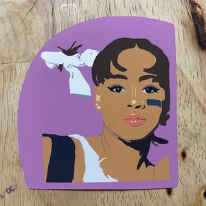 May include: A sticker featuring a digital illustration of a person with brown skin, black hair, and a white flower behind their head. The person is wearing a black and white shirt and has black eyeliner and a black stripe under their eye.