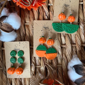 May include: Three pairs of handmade polymer clay earrings featuring pumpkin and leaf designs. The earrings are displayed on cards with the text "The Speckled Eye".