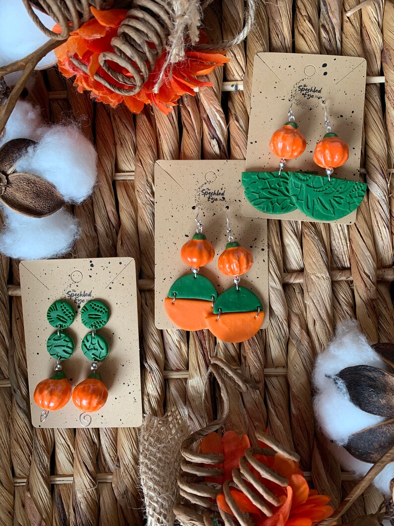 May include: Three pairs of handmade polymer clay earrings featuring pumpkin and leaf designs. The earrings are displayed on cards with the text "The Speckled Eye".