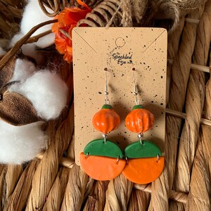 May include: A pair of dangle earrings featuring a green and orange pumpkin design. The earrings are on a brown card with the text "The Speckled Eye".