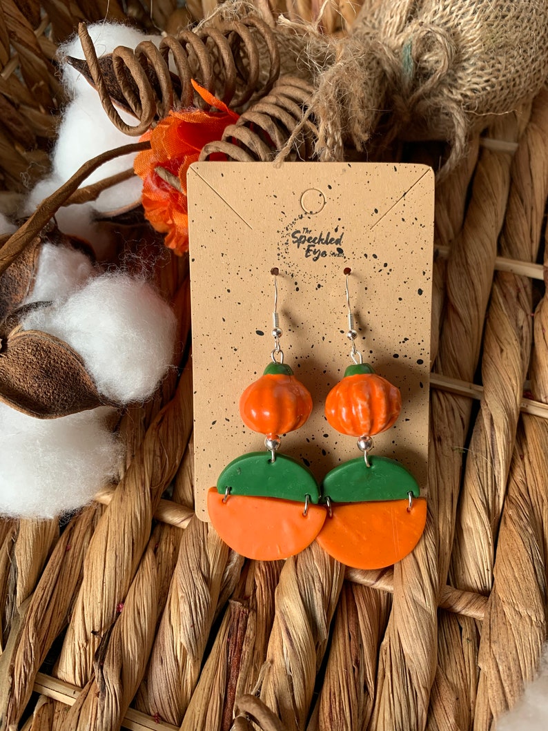 May include: A pair of dangle earrings featuring a green and orange pumpkin design. The earrings are on a brown card with the text "The Speckled Eye".