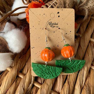 May include: A pair of pumpkin-shaped earrings with green leaf-shaped accents. The earrings are made of polymer clay and have a rustic, handmade look. The earrings are displayed on a brown card with the text "The Speckled Eye Shop" printed on it.