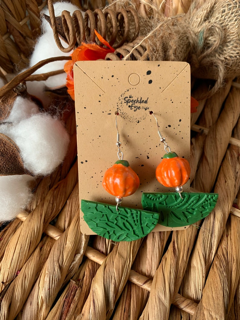 May include: A pair of pumpkin-shaped earrings with green leaf-shaped accents. The earrings are made of polymer clay and have a rustic, handmade look. The earrings are displayed on a brown card with the text "The Speckled Eye Shop" printed on it.