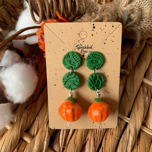 May include: A pair of dangle earrings featuring two green and white patterned clay circles and a small orange pumpkin charm. The earrings are displayed on a card with the text "The Speckled Eye Studio".