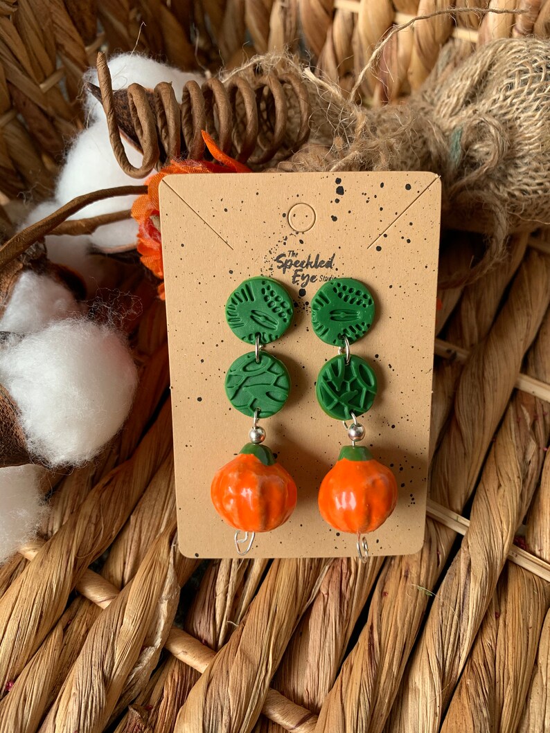 May include: A pair of dangle earrings featuring two green and white patterned clay circles and a small orange pumpkin charm. The earrings are displayed on a card with the text "The Speckled Eye Studio".