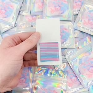 May include: A hand holding a small white card with a colorful, striped square of cotton candy-colored putty. The background shows many small, clear, iridescent bags with round stickers that read "Cotton Candy Clouds DOT DOT PUTTY" and a butterfly graphic.