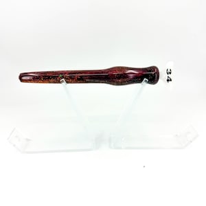 May include: A dark burgundy and black resin pen blank with embedded gold and iridescent glitter particles. The cylindrical object features a tapered shape and a small white tag attached to the end with the number 34 printed in black ink.
