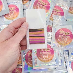 May include: A square piece of clay featuring horizontal stripes in black, orange, and magenta, attached to a white backing card. The background consists of multiple iridescent plastic pouches, each labeled with a circular sticker reading Muse Dot Dot Putty.