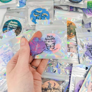 May include: A collection of small, holographic resealable pouches containing colourful putty, each labelled with a circular sticker featuring a butterfly icon and names like Pixie Sparkle, Sunken Treasure, and Witching Hour, with one purple and blue putty piece held in front.