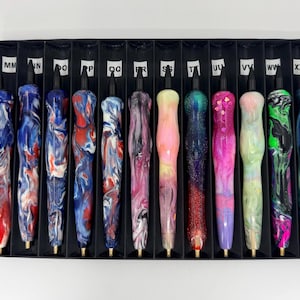 May include: A collection of twelve resin pens with varied marbled, glittered, and swirling color patterns, each featuring a black tip and end, arranged in a black partitioned display tray with white labels marked MM through XX at the top.