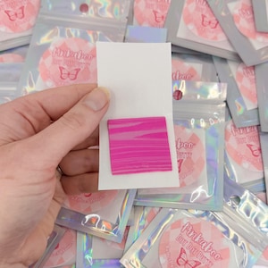 May include: A square piece of bright pink putty with a horizontal striped texture, attached to a white rectangular card. The background consists of multiple holographic resealable pouches, each featuring a circular label with a butterfly graphic.