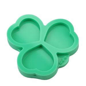 May include: A vibrant green silicone mould in the shape of a three-leaf clover, with each leaf formed as a heart. This mould is designed for crafting or baking, featuring a smooth surface and rounded edges. It is set against a white backdrop.