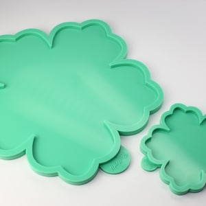 Puede incluir: Dos moldes de silicona verde con forma de trébol de cuatro hojas. El molde más grande tiene un diámetro de aproximadamente 18 cm y el más pequeño de aproximadamente 10 cm.