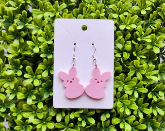 May include: A pair of pink bunny-shaped earrings with silver hooks. Each earring features a bunny silhouette with three small red dots for facial features. The earrings are displayed on a white card against a backdrop of green foliage.
