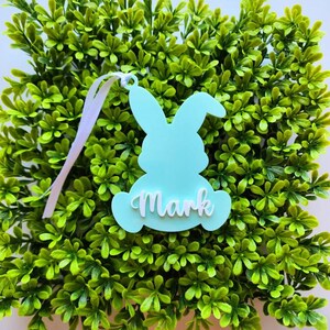 May include: A light blue bunny-shaped Easter tag with the name "Mark" written in white. The tag is hanging from a white ribbon and is surrounded by green foliage.