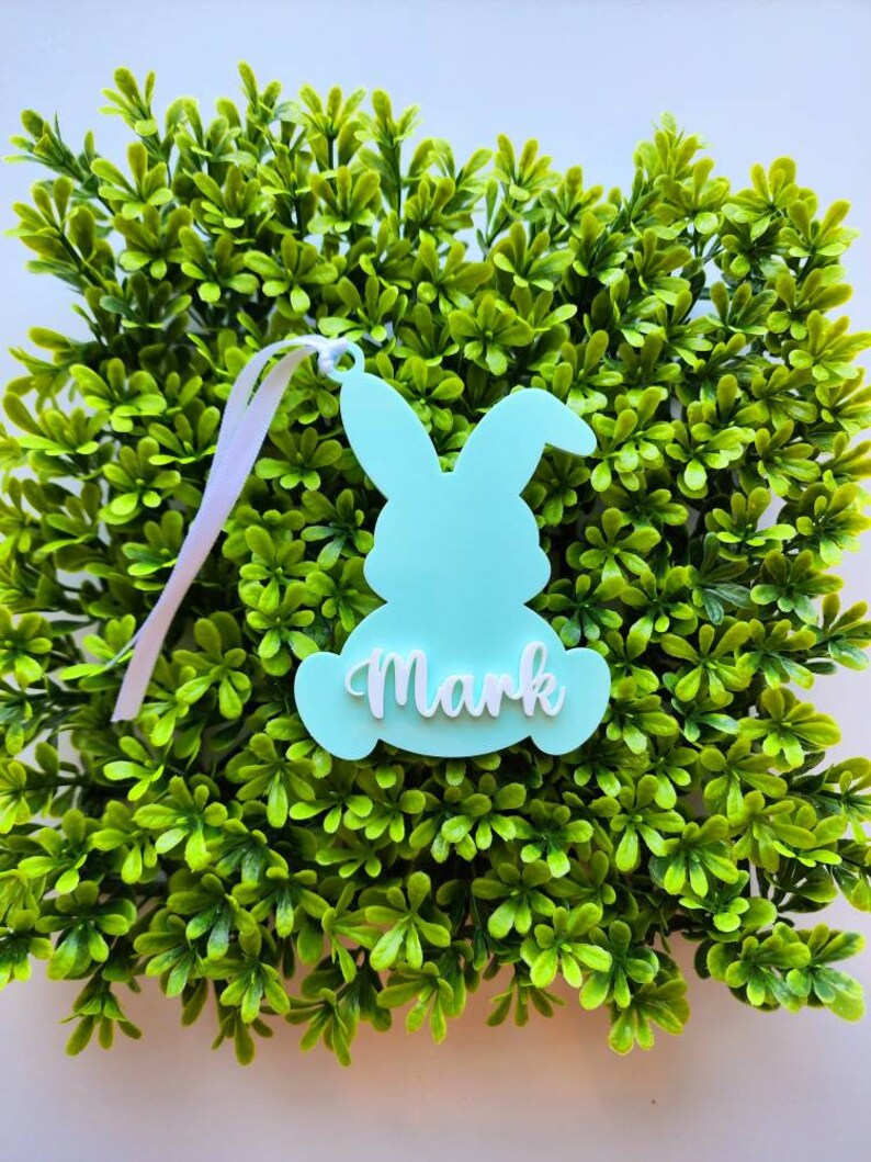 May include: A light blue bunny-shaped Easter tag with the name "Mark" written in white. The tag is hanging from a white ribbon and is surrounded by green foliage.