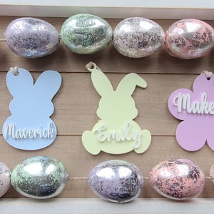 May include: A garland of pastel-colored Easter eggs with personalized bunny and egg-shaped ornaments. The ornaments are decorated with the names Julia, Maverick, Emily, Mackenzie, and Sofia.