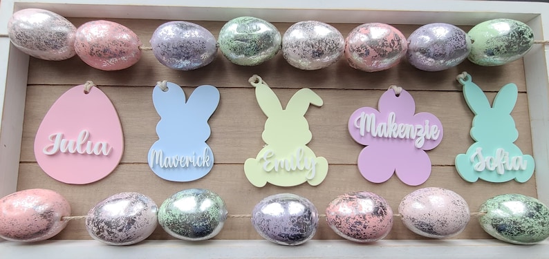 May include: A garland of pastel-colored Easter eggs with personalized bunny and egg-shaped ornaments. The ornaments are decorated with the names Julia, Maverick, Emily, Mackenzie, and Sofia.