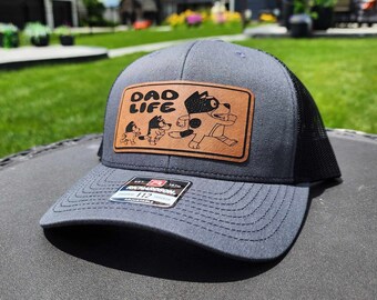 May include: A gray and black trucker hat with a brown leather patch that says "Dad Life" and features a cartoon dog. The patch also has three smaller cartoon dogs.