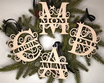 May include: Four wooden Christmas ornaments with intricate designs and the names "Corbin", "Mom", "Dad", and "Aalaya" cut out. The ornaments are hanging from black ribbon on a green pine branch.