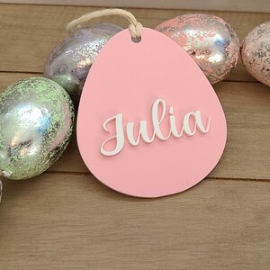 May include: Pink egg-shaped wooden tag with the name "Julia" written in white raised letters. The tag is surrounded by a garland of iridescent pastel Easter eggs.