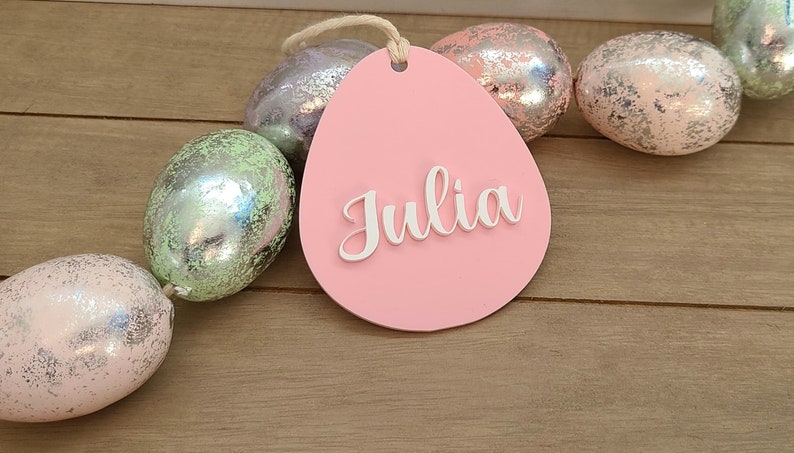 May include: Pink egg-shaped wooden tag with the name "Julia" written in white raised letters. The tag is surrounded by a garland of iridescent pastel Easter eggs.