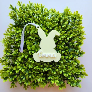 May include: A yellow bunny-shaped Easter ornament with the name "Emma" written on it. The ornament has a white ribbon attached to it.