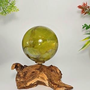 May include: A translucent olive green sphere with internal fractures and a smooth, polished surface, resting securely on a natural, irregularly shaped piece of brown driftwood that serves as a sturdy base for the decorative orb display.