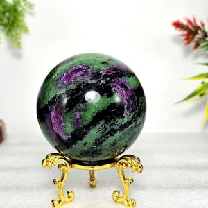 May include: A polished gemstone sphere with green, purple, and black marbling, displayed on a small, ornate gold-colored stand. The sphere's colors create a striking visual contrast, highlighting its natural beauty. The stand adds an elegant touch.