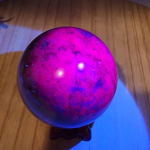 May include: A spherical stone orb displaying a vibrant, glowing magenta and deep purple mottled pattern under ultraviolet light. The polished surface reflects a bright light source, highlighting the smooth, rounded texture of the mineral sphere.